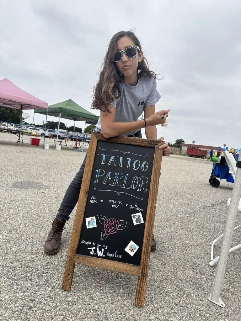 Woman leaning on a Tattoo Parlor chalkboard sign at an outdoor event
